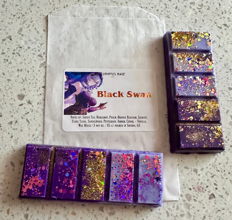 Puede incluir: Barra de cera fundida p&uacute;rpura y dorada con purpurina. La etiqueta dice "Black Swan" y enumera las notas de fragancia: T&eacute; verde, bergamota, melocot&oacute;n, flor de azahar, jazm&iacute;n, ylang ylang, s&aacute;ndalo, petitgrain, &aacute;mbar, cedro, vainilla. Cera fundida (85 g) vertida en Smyrna, GA.