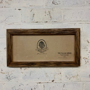 May include: A brown wooden picture frame with a brown paper insert. The insert has a black and white logo for "Royal Oak" and the text "The Timber Shack Hand Crafted Reclaimed Timber Furniture & Picture Frames Stony"