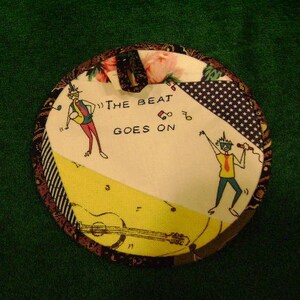 May include: A round, colorful fabric patch with a white background. The text "The Beat Goes On" is printed in black. There are two cartoon figures on the patch, one playing a guitar and the other holding a microphone.