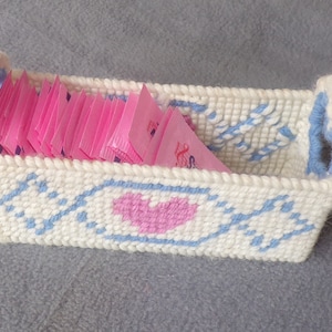 May include: A white crocheted basket with a blue and white geometric pattern and a pink heart design. The basket is filled with pink sugar packets.