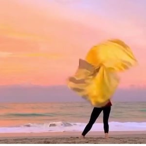May include: A person holding a yellow fabric in the air, with the ocean and a pink and orange sunset in the background.