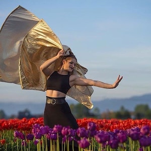 Puede incluir: Una mujer con un top corto y una falda negros baila en un campo de tulipanes rojos y morados, sosteniendo una bandera de tela dorada.