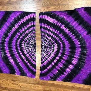May include: Two tie-dye fabric pieces with a spiral design in black, purple, and white. The fabric is laid out on a wooden surface. The spiral pattern radiates from the center of each piece.