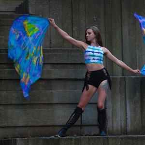 K&ouml;nnte beinhalten: Eine Frau tr&auml;gt ein tie-dye Crop-Top und schwarze Shorts mit Fransen und h&auml;lt zwei blaue, gelbe und violette Batik-Stofffahnen.