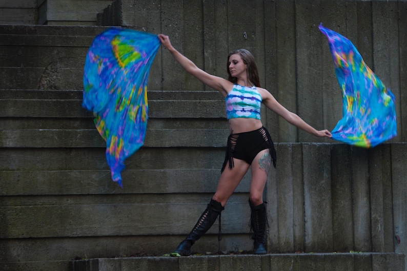 K&ouml;nnte beinhalten: Eine Frau tr&auml;gt ein tie-dye Crop-Top und schwarze Shorts mit Fransen und h&auml;lt zwei blaue, gelbe und violette Batik-Stofffahnen.