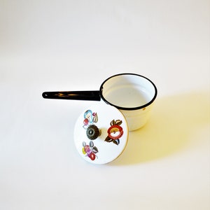 May include: A white enamel saucepan with a black handle and rim. The lid features a floral design with red, yellow, and blue accents. The saucepan is a vintage kitchenware item.
