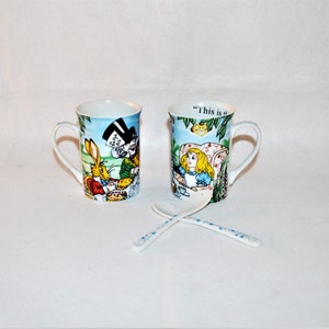 Könnte beinhalten: Zwei weiße Keramikbecher mit Illustrationen aus Alice im Wunderland. Ein Becher zeigt den Hutmacher und das weiße Kaninchen bei einer Teeparty. Der andere Becher zeigt Alice. Zwei weiße Löffel mit blauen Blumenmustern sind ebenfalls vorhanden.