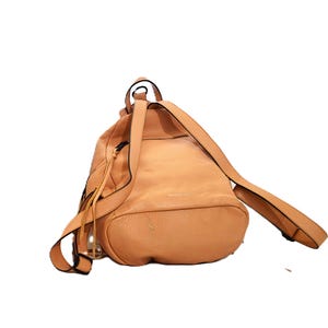 May include: A light brown leather backpack with adjustable straps. The bag has a rounded shape with a drawstring closure and a small handle at the top. The straps are thin and the bag appears to be empty.