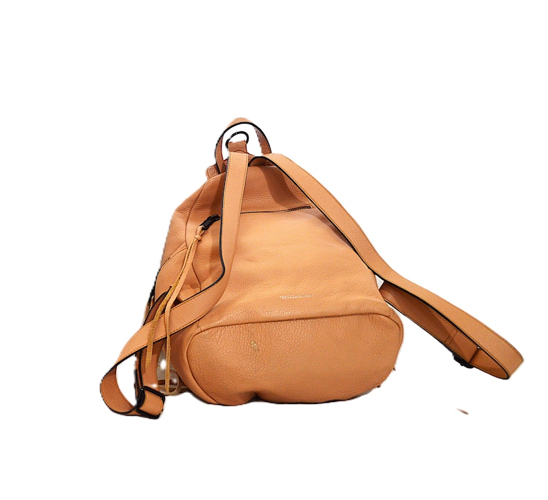 May include: A light brown leather backpack with adjustable straps. The bag has a rounded shape with a drawstring closure and a small handle at the top. The straps are thin and the bag appears to be empty.