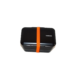 May include: A rectangular black bento box with rounded corners and a smooth finish. The container is secured by a bright orange elastic band wrapped vertically around the center. The word TAKENAKA is printed in white on the top lid surface.