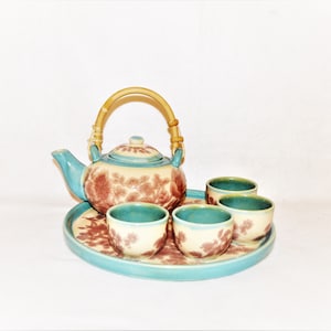 Puede incluir: Un juego de té de cerámica que incluye una tetera con asa de bambú y tres tazas a juego dispuestas sobre una bandeja redonda. Las piezas son de color crema con bordes verde azulado y patrones florales marrones en la superficie.