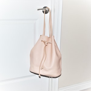 May include: A blush-colored leather bucket bag with long straps, hanging from a silver door handle. The bag has a drawstring closure and a rounded shape, suggesting a casual style. The bag's soft color and design make it suitable for everyday use.