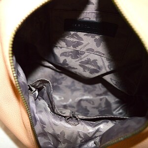 May include: Close-up of a peach-colored handbag with a zipper closure. The interior features a gray lining with a butterfly pattern and a black label with the brand name. The bag's design is simple and elegant.