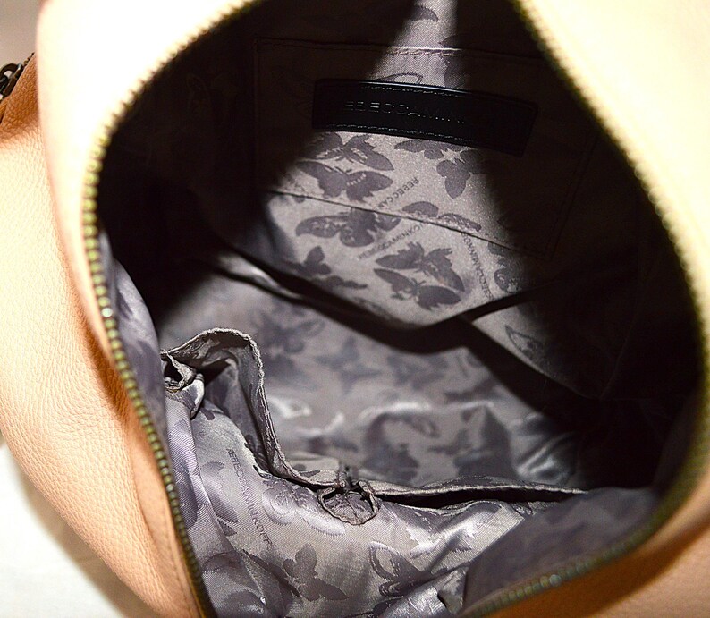 May include: Close-up of a peach-colored handbag with a zipper closure. The interior features a gray lining with a butterfly pattern and a black label with the brand name. The bag's design is simple and elegant.