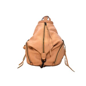 May include: A light brown leather backpack with multiple zippered compartments and a top handle. The bag has a rounded shape and features black zippers and hardware. The design includes long zipper pulls and a black clasp.