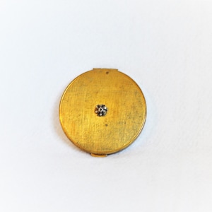 May include: A round, antique gold-toned compact with a small, silver-colored floral embellishment in the center. The compact is open, revealing a mirror and powder compartment. The compact is on a white background.