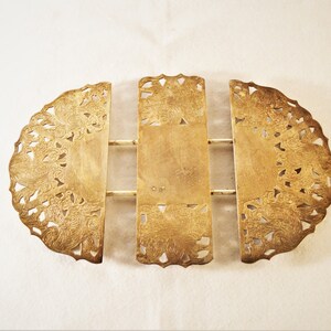 May include: An expandable gold-toned metal trivet featuring three sections connected by sliding bars. The two outer semi-circular pieces and the central rectangular piece display intricate, ornate cut-out patterns with detailed floral and scroll motifs.