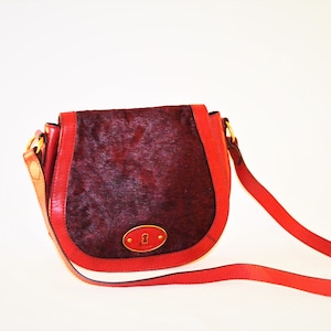 May include: A saddle-shaped crossbody bag featuring a dark burgundy textured hair-on-hide front flap and smooth red leather trim. The bag includes a gold-toned oval metal plate with a keyhole cutout on the front and a matching red leather strap.