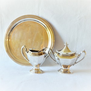 May include: A silver tea set with a round tray, a creamer, and a sugar bowl. The tray is large and circular. The creamer and sugar bowl have a geometric design and handles. The set is on a white background.