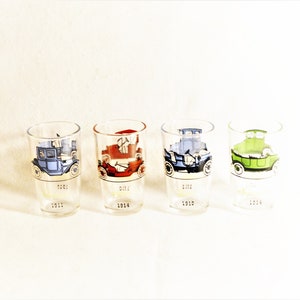 May include: Four clear shot glasses with vintage car designs in blue, red, blue, and green. Each glass features a different car model and the year it was made: 1911, 1914, 1910, and 1914. The glasses are arranged in a row.