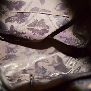 May include: Interior view of a dark-colored handbag. The lining features a repeating butterfly pattern in shades of gray and purple. The brand name "Rebecca Minkoff" is visible on the lining.
