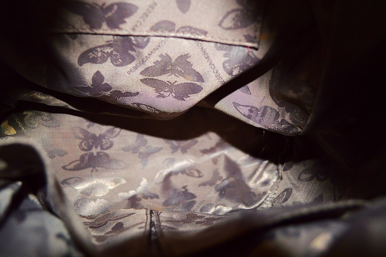 May include: Interior view of a dark-colored handbag. The lining features a repeating butterfly pattern in shades of gray and purple. The brand name "Rebecca Minkoff" is visible on the lining.