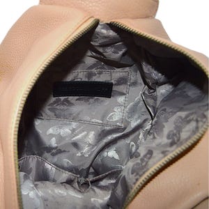 May include: A light pink leather handbag with a zipper closure. The interior features a gray lining with a butterfly pattern and a black label that reads "REBECCA MINKOFF". The bag is open, revealing the inside.