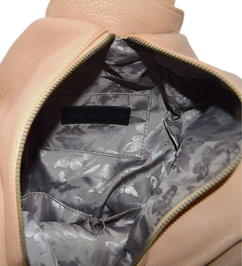 May include: A light pink leather handbag with a zipper closure. The interior features a gray lining with a butterfly pattern and a black label that reads "REBECCA MINKOFF". The bag is open, revealing the inside.