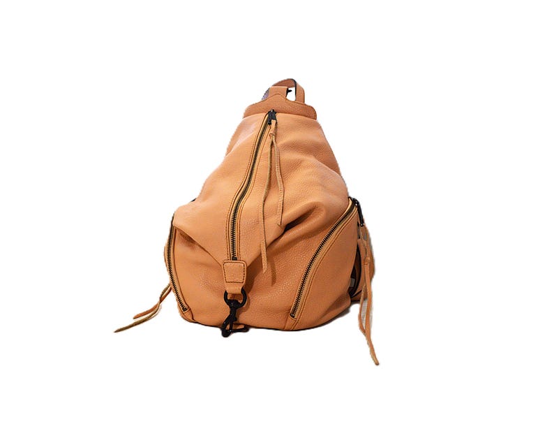 May include: A tan leather backpack with multiple zippered compartments and black hardware. The bag has a rounded shape and features long zipper pulls and a top handle. The leather has a textured appearance.