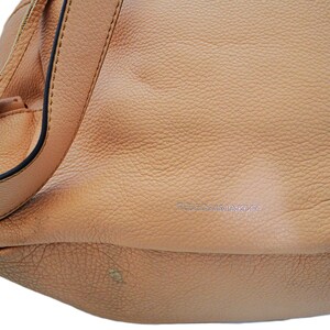 May include: A light tan leather handbag with a textured surface. The bag has a shoulder strap with fringe detailing and a black trim. The brand name is subtly embossed on the front of the bag.