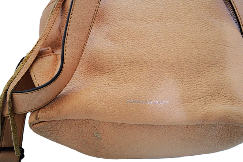 May include: A light tan leather handbag with a textured surface. The bag has a shoulder strap with fringe detailing and a black trim. The brand name is subtly embossed on the front of the bag.