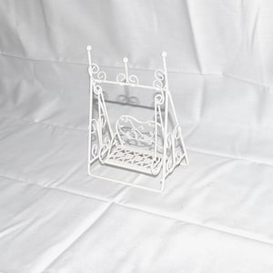 May include: A white metal decorative swing with a heart-shaped back and a latticed seat. The swing has a delicate, ornate design with scrollwork details. The swing is a miniature decorative item.