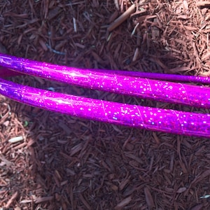 May include: Two purple glitter hula hoops with a spiral pattern. The hoops are lying on a bed of brown wood chips.