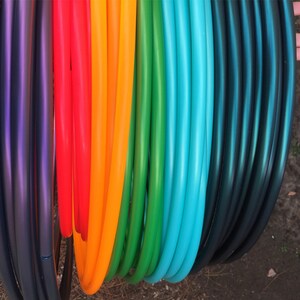 May include: A selection of brightly coloured hula hoops, arranged in a rainbow gradient. The hoops are displayed vertically, with shades of purple, red, orange, green, blue, and teal. The smooth, curved tubes are made of plastic.