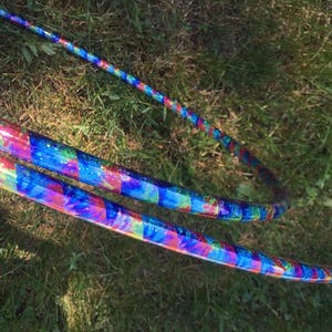May include: Two colourful iridescent hoops, one on top of the other, with a blue, pink and green pattern. The hoops are lying on a bed of green grass.