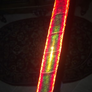 May include: A close-up of red and green reflective tape wrapped around a cylindrical object. The tape has a geometric pattern and is illuminated by a bright light.