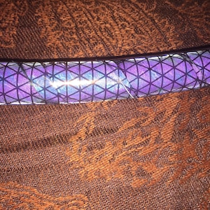 May include: A close-up of a purple and black iridescent hoop with a geometric pattern. The hoop is lying on a brown fabric surface.