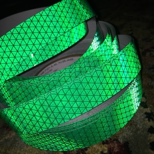 May include: A roll of vibrant green reflective tape featuring a repeating triangular design. The tape is coiled, showing the white backing. The surface of the tape has a shiny, textured finish, reflecting light.