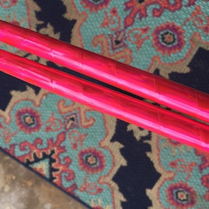 May include: Two pink iridescent glow sticks, lying on a patterned rug.