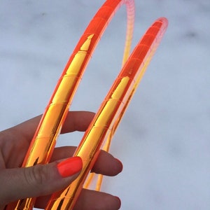 May include: Two orange and gold metallic hula hoops. The hoops are held in a hand with bright orange nail polish.