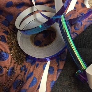 May include: A roll of iridescent blue and green tape with a white backing. The tape is wrapped around a spool and is partially unwound. The tape is lying on a brown fabric with blue swirls.