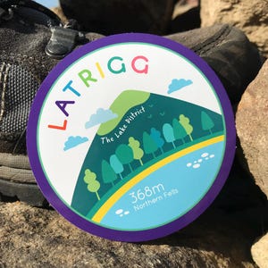 May include: A purple, white, and green round sticker with the text "LATRIGG" in rainbow colors. The sticker features a cartoon illustration of a green hill with trees and a blue lake. The text "The Lake District" and "368m Northern Fells" are also included on the sticker.