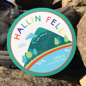 May include: A round sticker with a green background and a white outline. The sticker features a colorful illustration of a mountain with the text "Hallin Fell" in a rainbow arc above the mountain. The text "The Lake District" is below the mountain. The text "388m Far Eastern Fells" is at the bottom of the sticker.