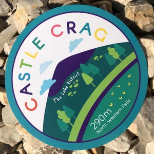 May include: Round sticker with a teal border and the words "CASTLE CRAG" in colorful letters. The design includes a mountain scene with trees, clouds, and the text "The Lake District" and "290m North Western Fells".