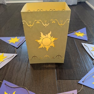 May include: A purple and yellow paper banner with a sun design. The banner is strung with twine and has a yellow paper lantern with a sun design in the background.