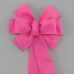 May include: A large pink bow made from a textured ribbon. The bow has a long tail and is tied in a classic style.