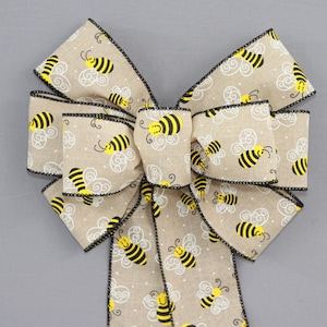 May include: A beige burlap bow with black trim and a yellow and black bee pattern. The bow is tied with a long tail and has a white polka dot pattern.