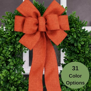 May include: A large, burnt orange fabric bow with two long, hanging tails centered on a circular green leaf wreath. A circular green badge on the lower right side features the text 31 Color Options in black lettering.