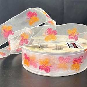 May include: Sheer white ribbon with a repeating pattern of orange and pink butterflies. The ribbon is rolled on a cardboard spool, with some ribbon unrolled and draped over the top. The butterflies have a glittery appearance.