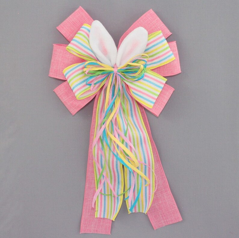 Op de afbeelding: Een roze en wit gestreepte strik met konijnenoren. De strik is versierd met pastelkleurige linten en is perfect voor Pasen.